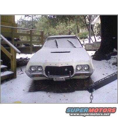 Front view of a white 1972 Ford Ranchero covered in snow, parked outside next to a wooden structure and a tree