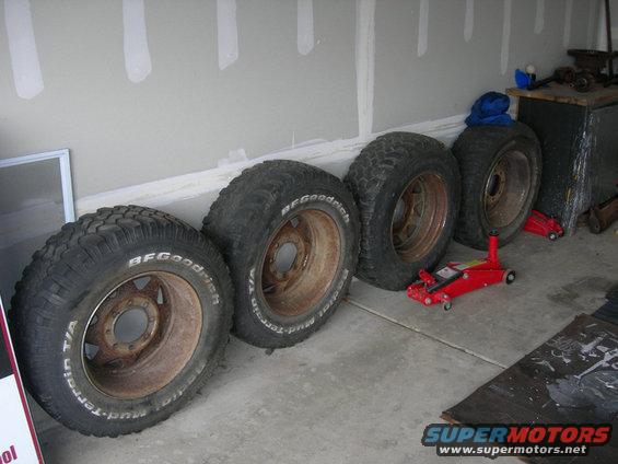 Four BFGoodrich Mud-Terrain T/A tires mounted on rusty steel wheels, with a red floor jack nearby