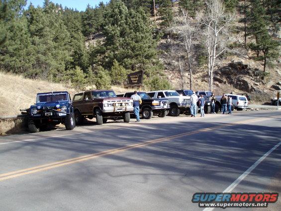 1979 Ford Bronco and other SUVs parked on roadside with trees and hills in background