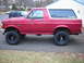 1995 Ford Bronco - A red Ford Bronco with a white removable top is parked on a driveway, showing its lifted suspension and off-road tires.