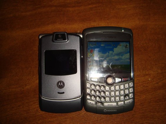 Two cell phones on a wooden surface. A Motorola RAZR is on the left, and a Blackberry is on the right