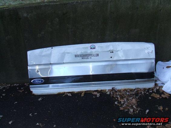 White Ford tailgate with a black stripe and chrome trim, dented and weathered