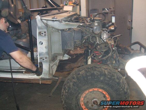 White Ford Bronco undergoing rebuild, front fender removed, engine exposed, large off-road tire visible