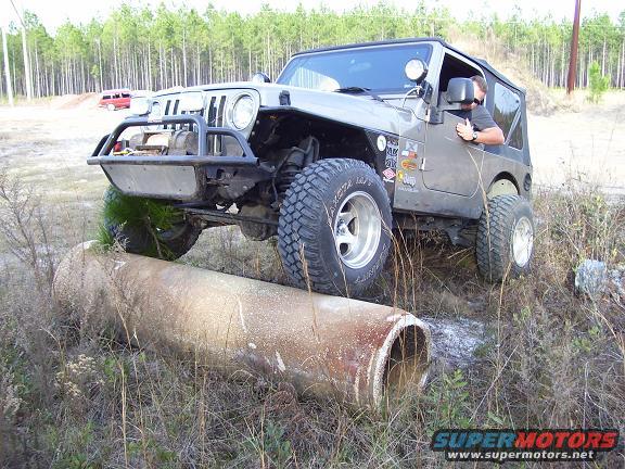 Gray Jeep TJ driving over a large pipe on a dirt path. Driver is visible
