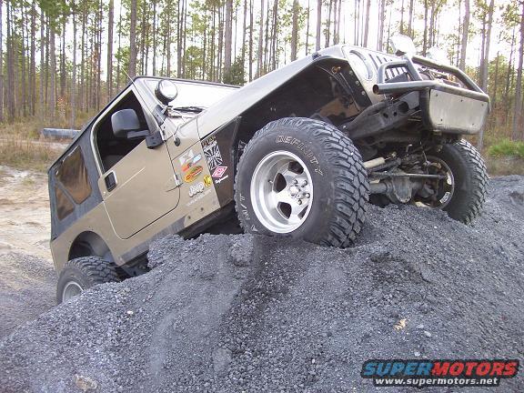 Tan Jeep Wrangler climbing a pile of gravel, front-right wheel elevated