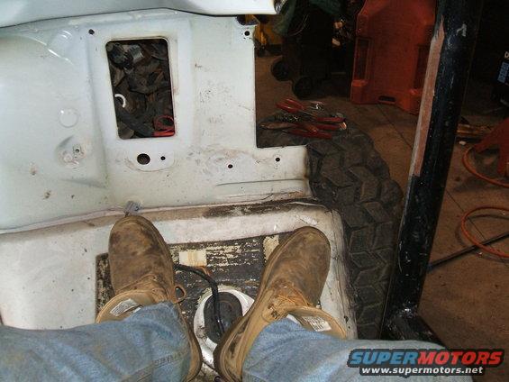 Interior of a white vehicle with exposed metal floor, a spare tire, and a person's legs wearing work boots