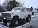 1983 Ford Bronco - A white Ford Bronco with oversized tires sits in a snowy driveway, with two vans visible in the background.