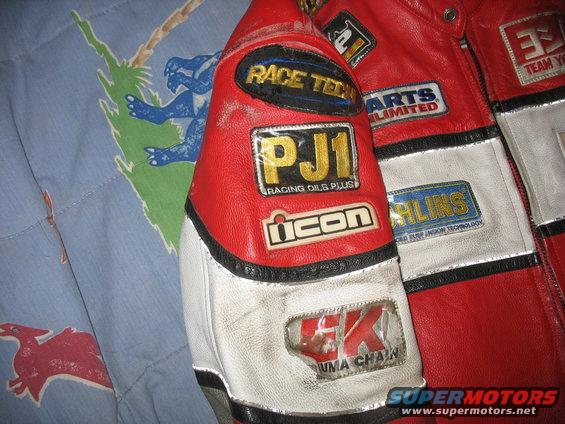 Red and white motorcycle jacket with various sponsor patches including PJ1, Icon, Race Tech, Ohlins, and EK