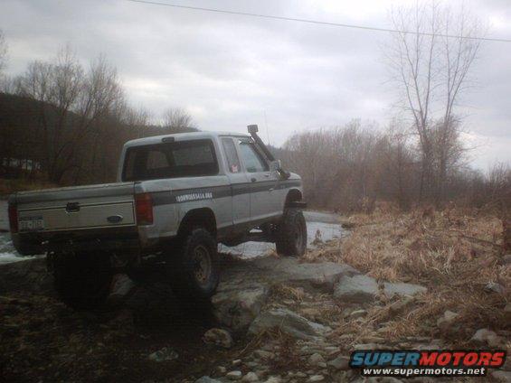 1990 Ford Ranger pickup truck with snorkel, driving over rocks in a muddy, wooded area