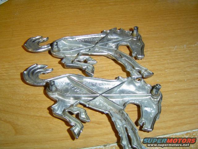 Two chrome horse emblems with mounting posts on a wooden surface. One emblem has visible part numbers