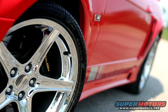 Close-up of a red car's chrome wheel with 'Saleen' written on the brake caliper. Silver racing stripes are on the side