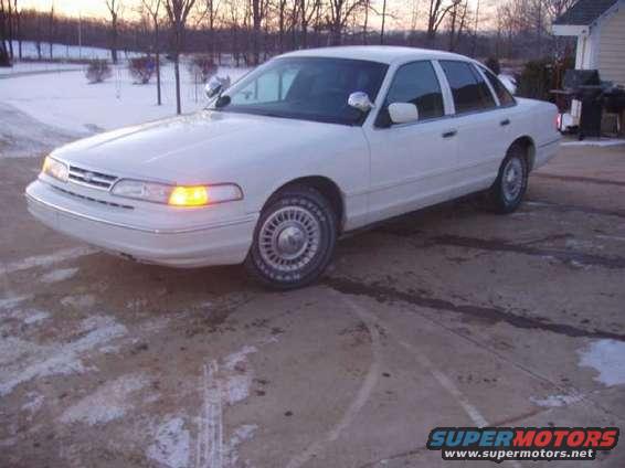 White sedan parked on a dirt lot with snow in the background. The car has a chrome grille and wire-spoke hubcaps