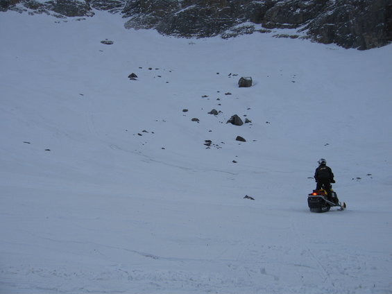 Person on a snowmobile on a wide, snow-covered slope with rocks and a mountain in the background