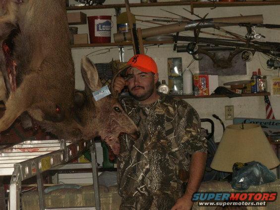 Man in camouflage holding a harvested deer, with shelves of hunting gear and supplies in the background