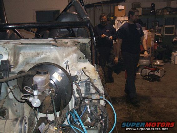 Vehicle chassis with exposed engine bay, brake booster, and wiring. Two men stand in the background