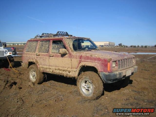 Mud-covered red Jeep Cherokee stuck in mud, being towed by a white Jeep
