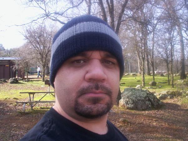 Man in beanie and black shirt in a wooded area with picnic tables and a building