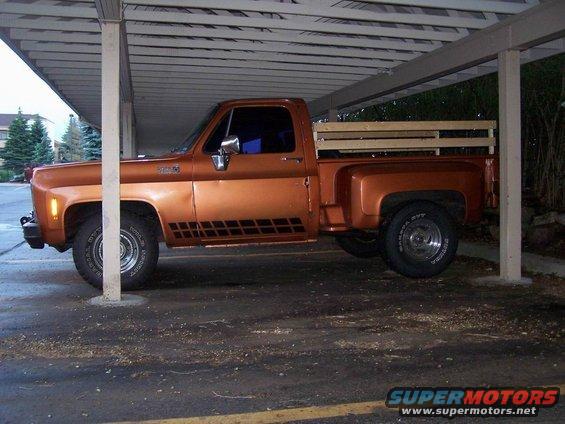 Orange pickup truck parked under a carport, side view. It has a wooden bed insert and black checkered decals on the door