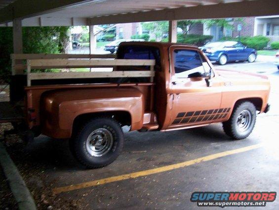 Orange pickup truck with wooden bed rails parked under a carport. A blue car is visible in the background
