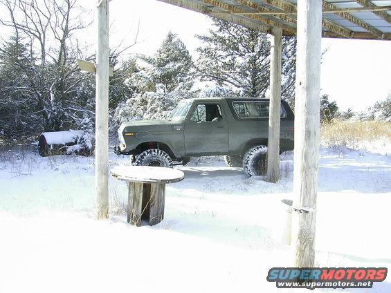 1978 Ford Bronco parked under a wooden shelter in a snowy landscape. The vehicle is dark green with off-road tires