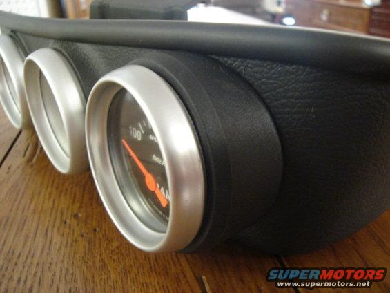 Gauge cluster with three gauges, one showing 100/100 and an orange needle. The housing is black and textured