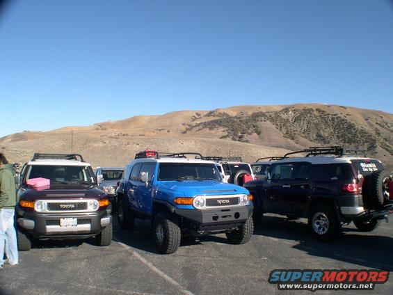 Three Toyota FJ Cruisers parked in a lot with hills in the background. The front FJ is blue, the middle is dark purple, and the one on the left is dark red