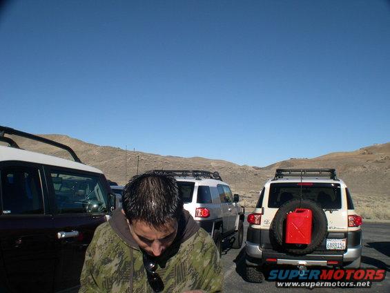 Two Toyota FJ Cruisers parked, one with a red gas can on the spare tire. Man in camo jacket in front