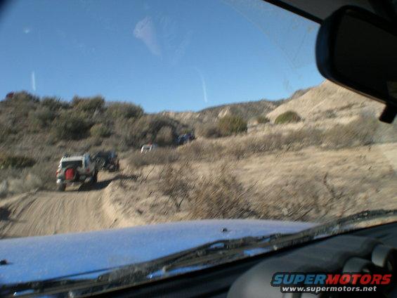 Several off-road vehicles driving on a dusty trail in a dry, hilly area