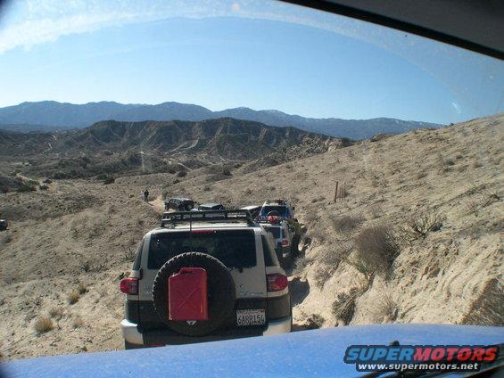 White Toyota FJ Cruiser with a red jerrycan on spare tire, driving on a dirt road in a desert landscape