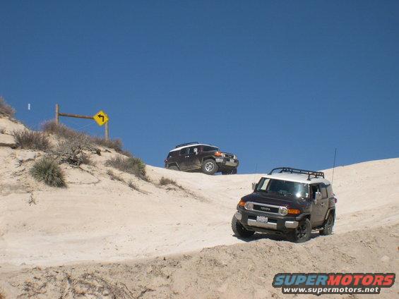 Two dark-colored 2007 Toyota FJ Cruisers driving on a sandy hill with a blue sky