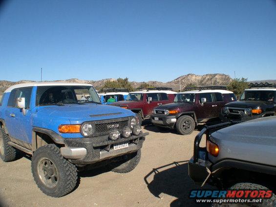 Several Toyota FJ Cruisers parked in a dirt lot with mountains in the background. One blue FJ is in the foreground