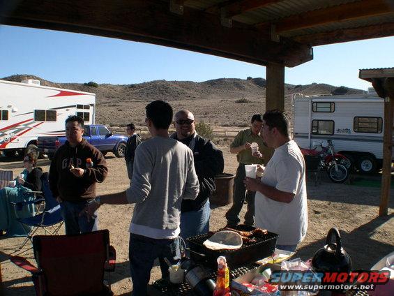 People gathered around a picnic table with a grill, RVs and a blue pickup truck in the background