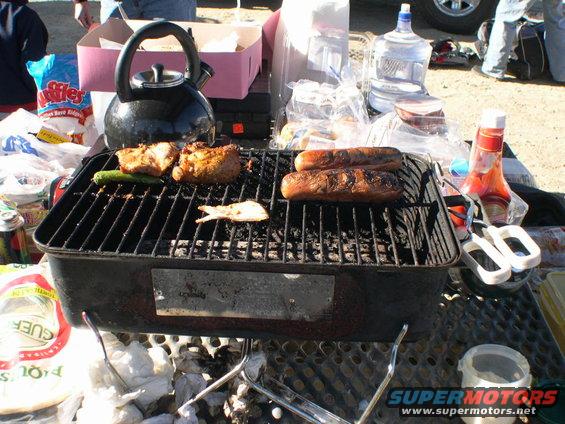 Portable grill with chicken and hot dogs cooking, kettle, drinks, and condiments on a table