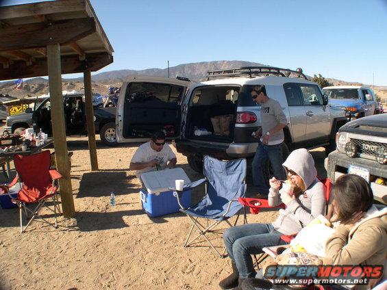 Two Toyota FJ Cruisers parked, one silver and one blue, with people gathered around a picnic table and chairs