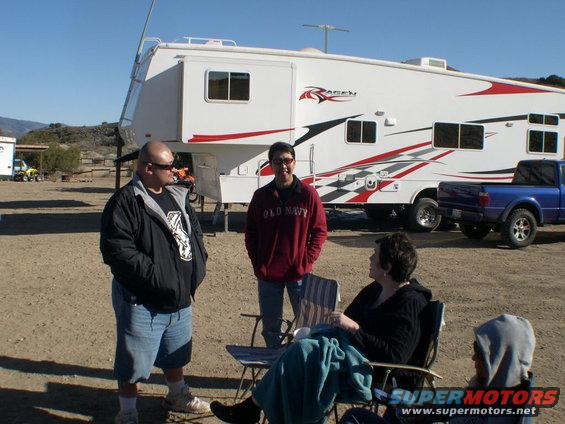 Three people stand and sit around a large RV with a blue pickup truck parked behind it