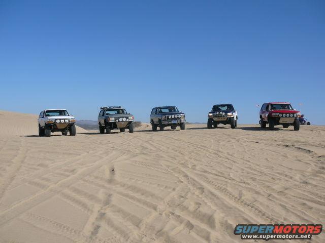 Five off-road SUVs parked on sand dunes under a clear blue sky