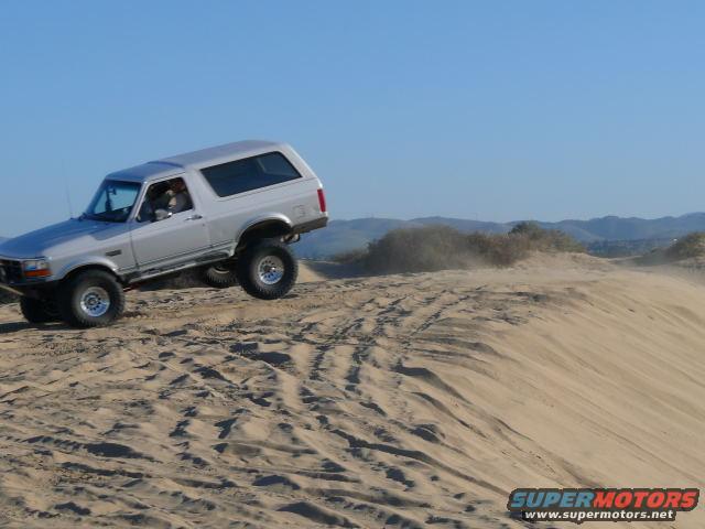 Silver SUV kicking up sand while driving on a dune. It has aftermarket wheels and appears to be lifted