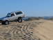 1988 Ford Bronco - A silver SUV navigates a sandy terrain, kicking up dust as it goes under a clear blue sky.