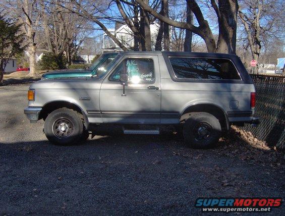 Silver 1992 Ford Bronco, side view, parked on gravel with a green truck behind it