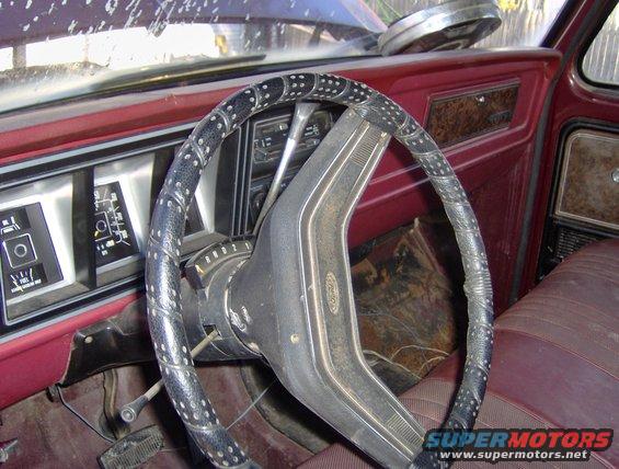 Interior shot of a vehicle's dashboard and steering wheel. The steering wheel is wrapped in black material with white dots. The dashboard has gauges and a red interior