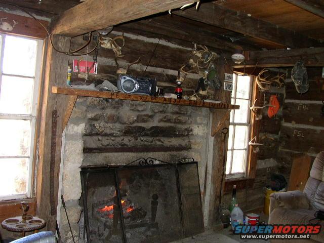 Interior of a rustic cabin with a stone fireplace, wooden beams, and animal antlers mounted on the walls. A radio sits on the mantelpiece