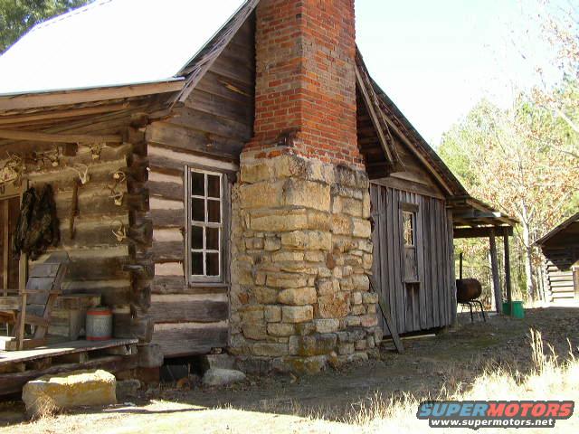 old log cabin with a large stone and brick chimney, porch with hanging pelts, and a wooden roof