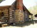 1995 Ford Bronco - A rustic log cabin with a prominent stone and brick chimney stands under a bright sky, surrounded by trees.
