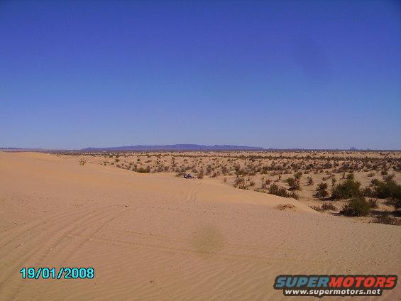 Desert dunes under a clear blue sky with a small vehicle in the distance. Date stamp 19/01/2008
