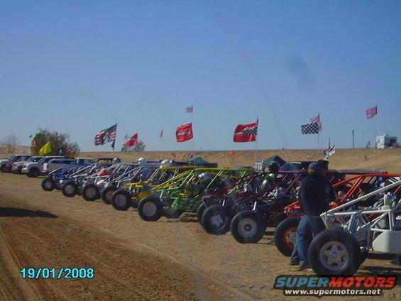 Line of colorful dune buggies parked on sand, with flags in the background