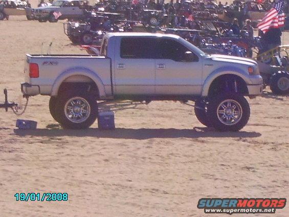 Silver lifted Ford F-150 pickup truck with custom wheels, parked on sand. Other vehicles and people in background