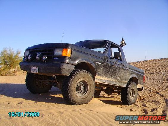 Black Ford Bronco with off-road tires parked on sand dunes. Has a pirate flag on the antenna
