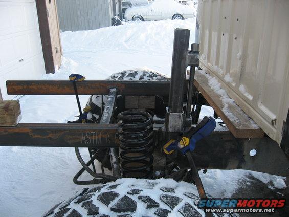 Truck frame with large coil spring and metal tubing, clamps attached. Snow on ground and tires