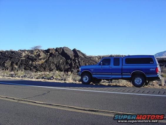 Blue pickup truck parked on the side of a road with a rocky hill in the background. It has a camper shell on the bed