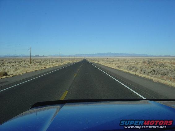 Blue truck hood in foreground, highway stretches to horizon in desert landscape